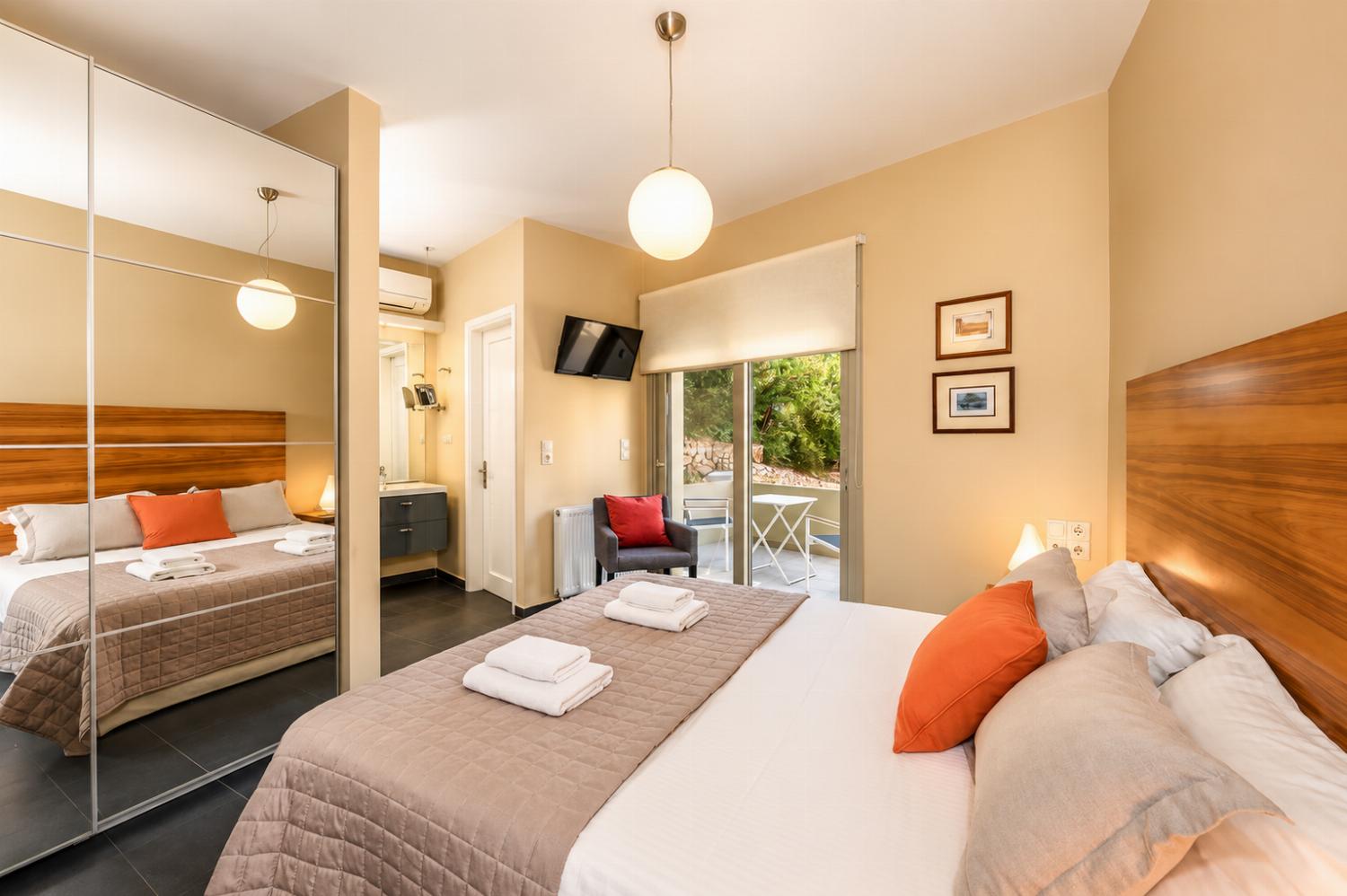 A bright bedroom features a double bed with a wooden headboard, a large mirrored wardrobe, and glass doors opening to a private outdoor seating area.