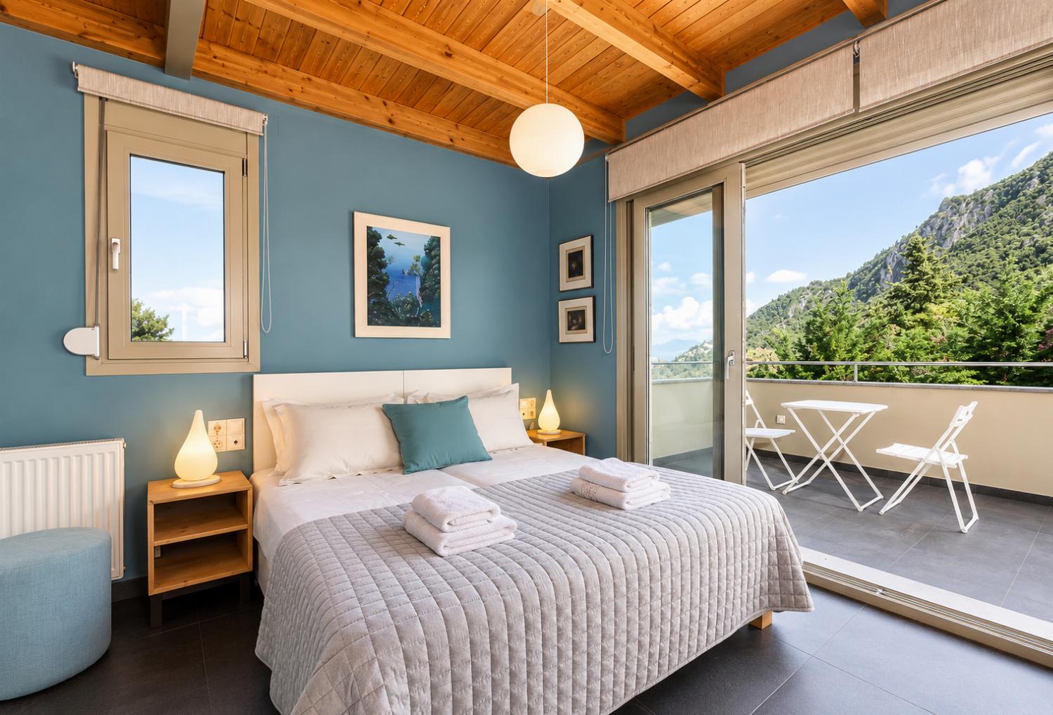 A charming bedroom features a double bed under an exposed wooden ceiling, blue accent walls, and wide glass doors that open to a private balcony with a mountain view.