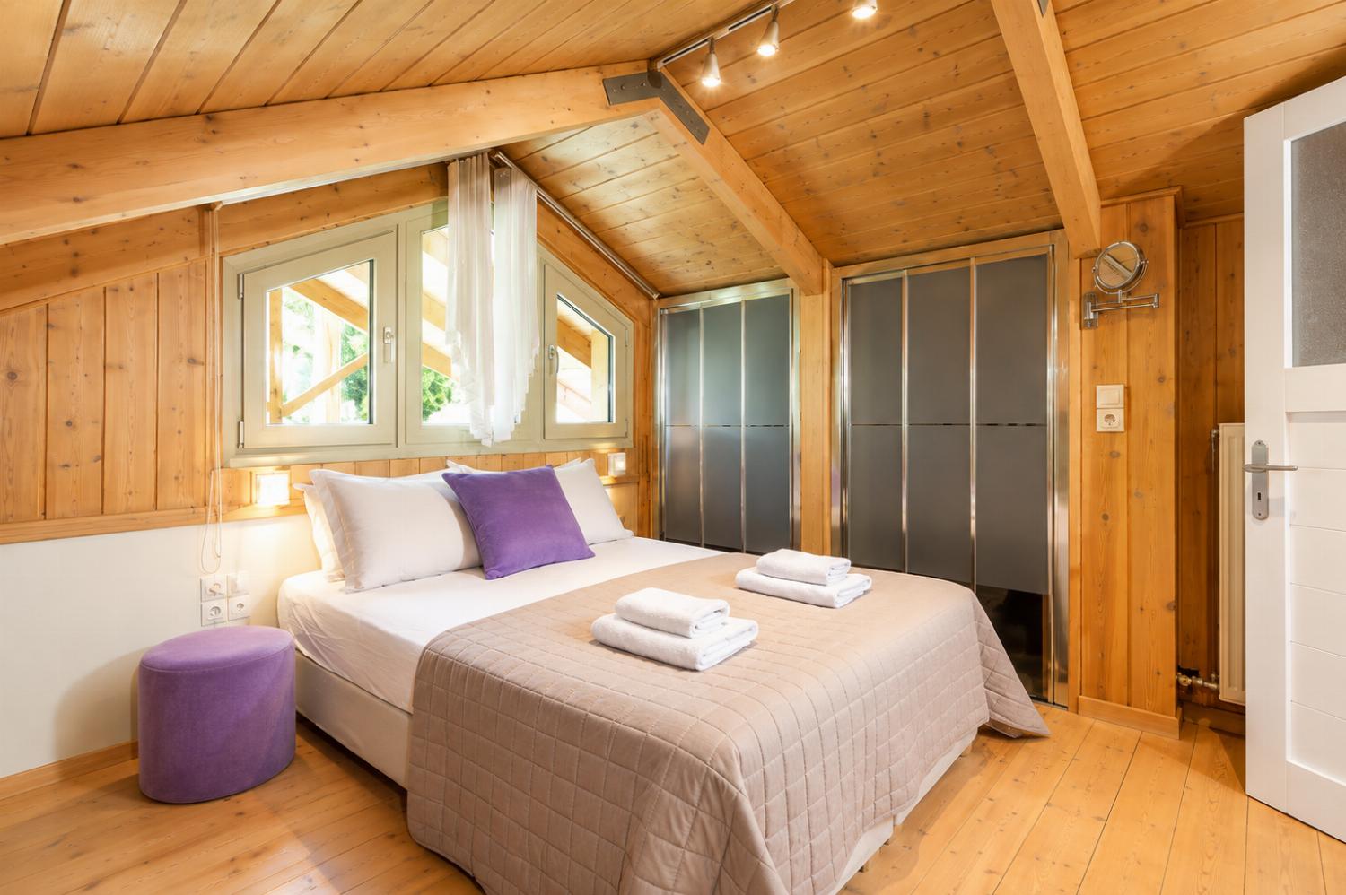 A rustic attic bedroom features wood-paneled walls and ceilings, a double bed with purple accent pillows, and tracking lights illuminating the sloped space.