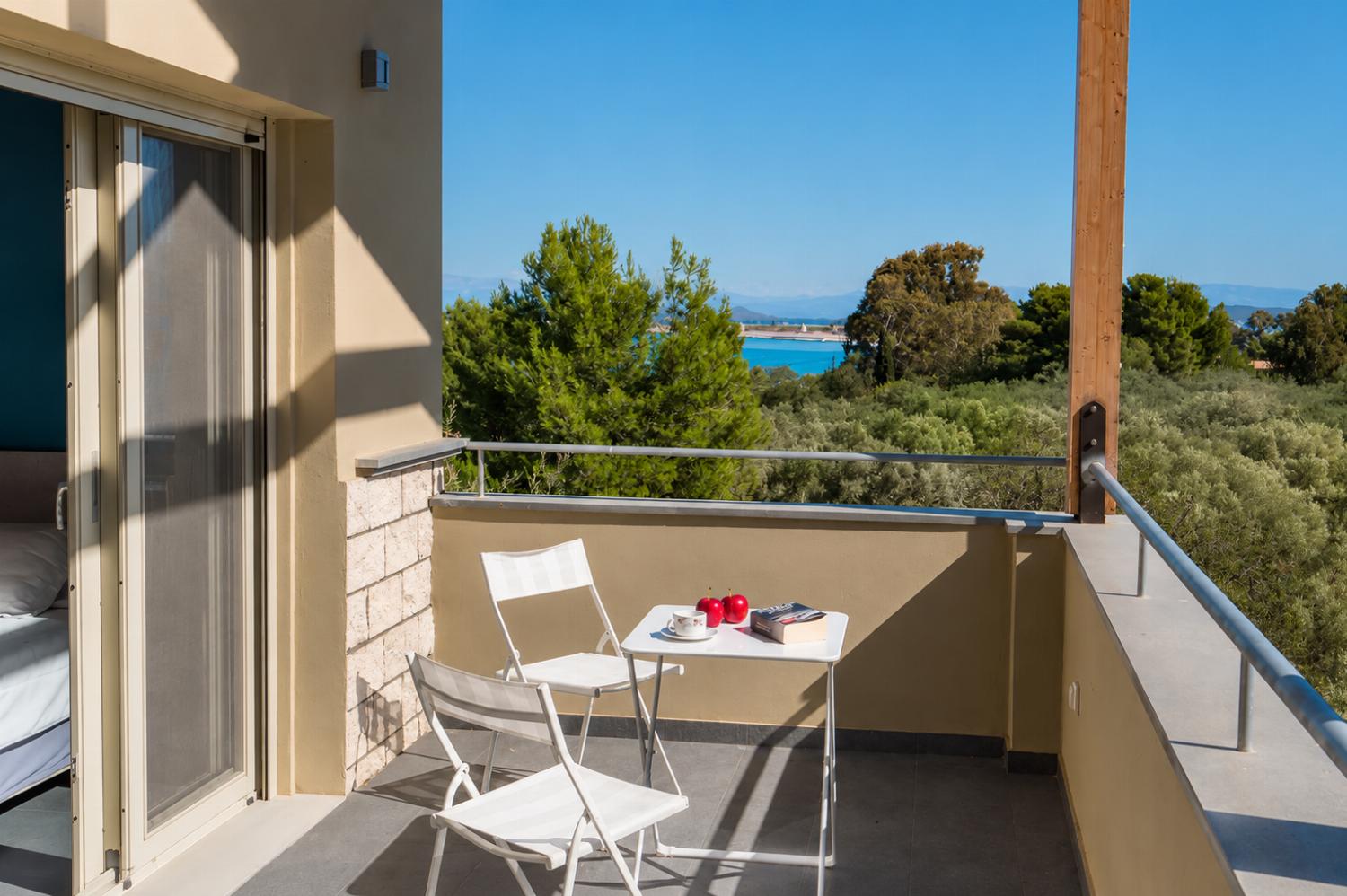 A sunlit balcony features a small white bistro table and two folding chairs, offering a scenic view of green treetops and the distant blue sea.