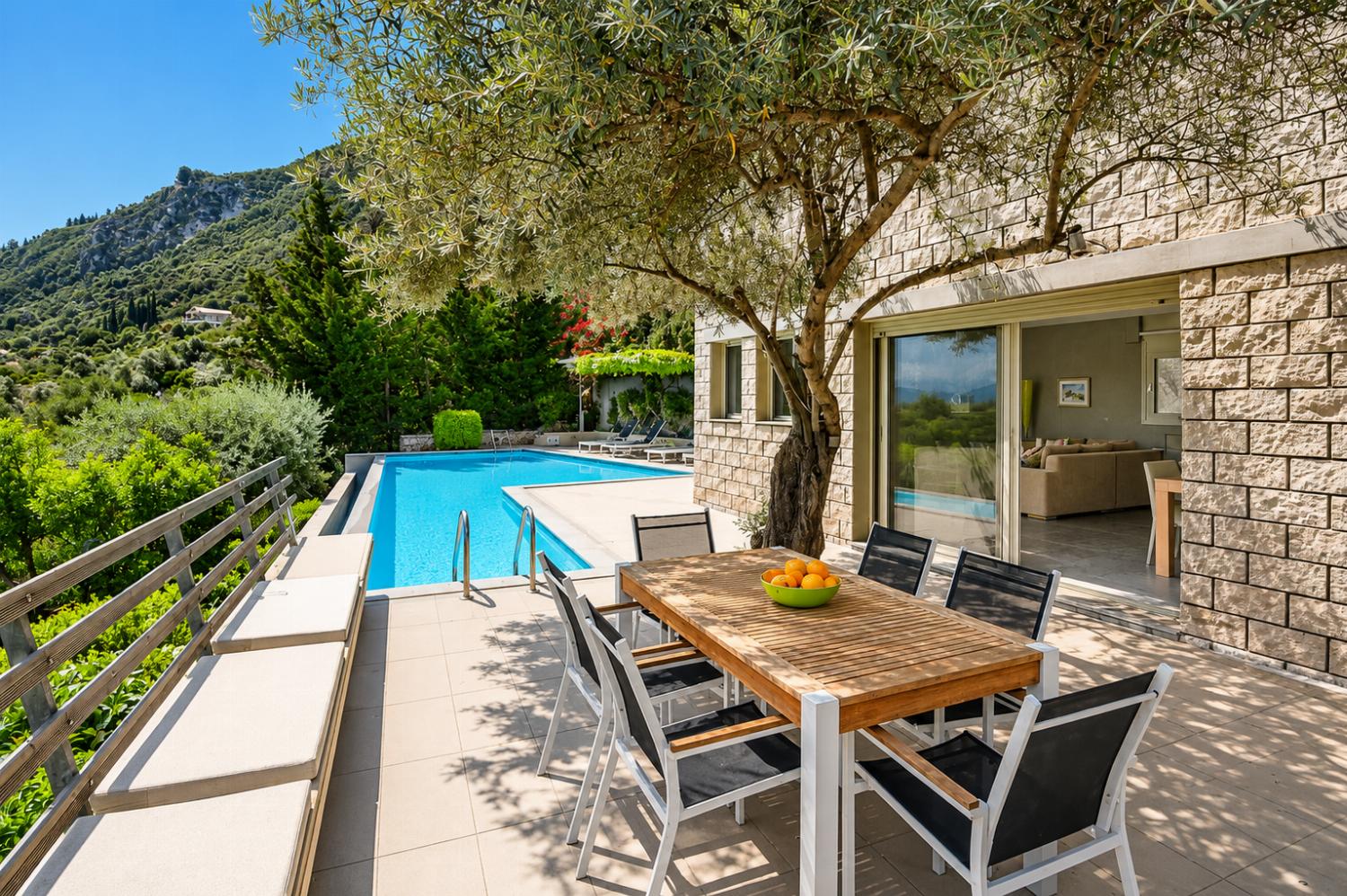 A wooden outdoor dining table sits on a sun-drenched terrace next to a stone villa, featuring a private swimming pool and a lush mountain landscape in the background.