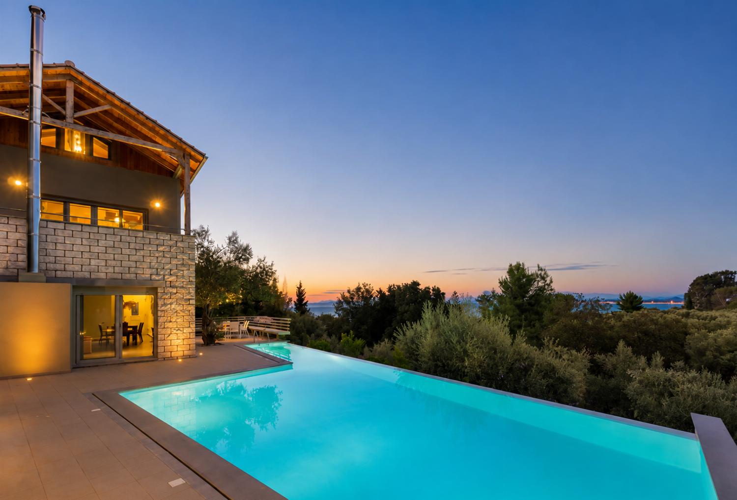 An illuminated outdoor swimming pool glows at dusk next to a multi-story stone villa, offering a panoramic view of the surrounding trees under a vibrant pink and purple sky.