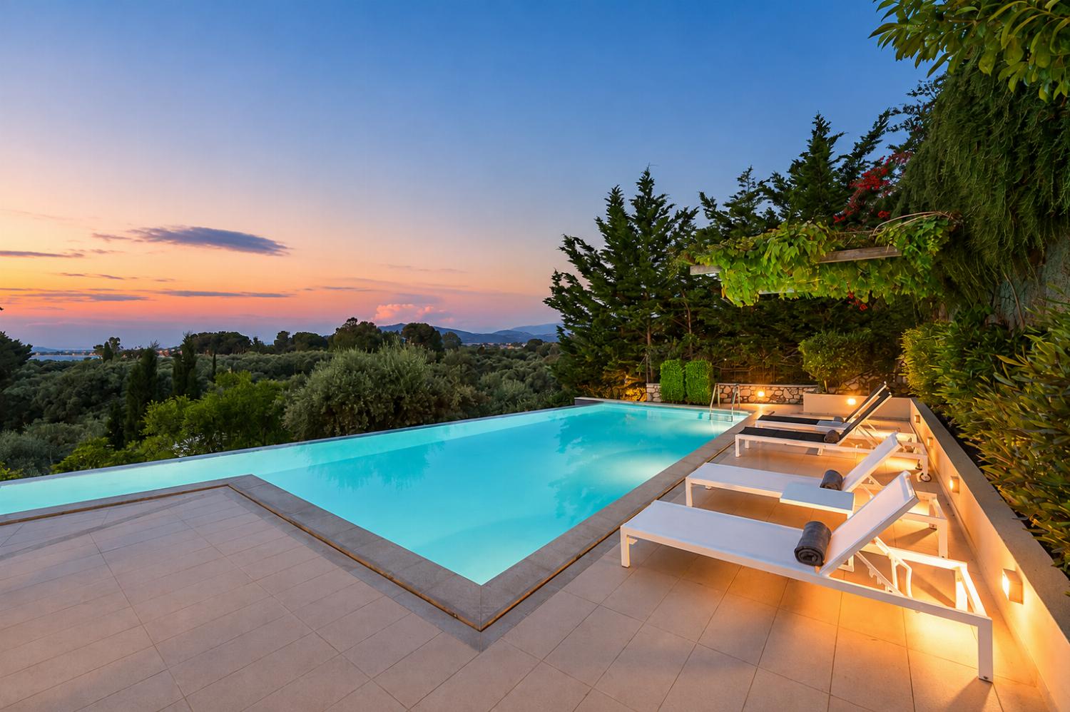 An illuminated outdoor swimming pool is lined with white lounge chairs at dusk, showcasing a scenic view of green treetops and mountains under a soft pink sky.