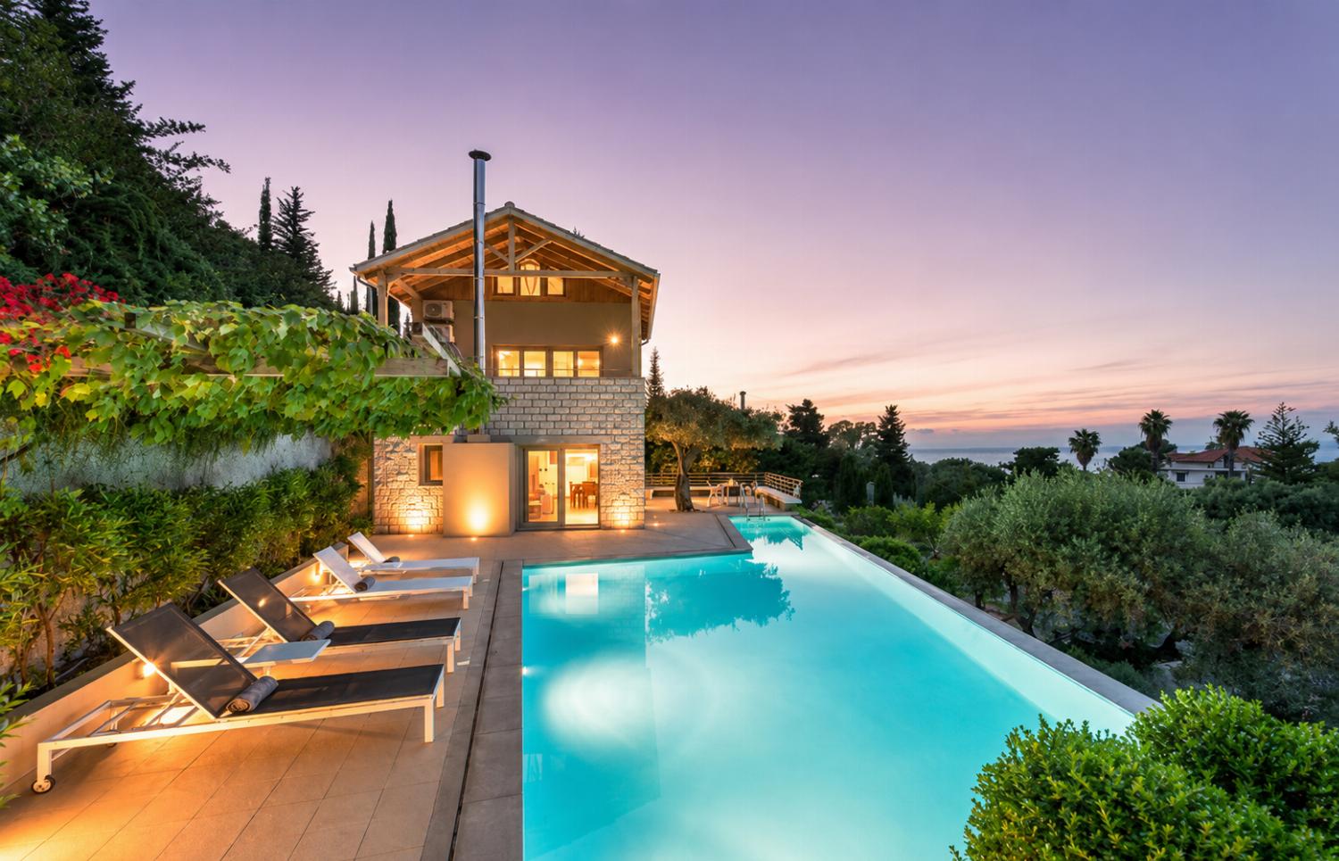 An illuminated infinity swimming pool highlights a multi-story stone villa at dusk, complete with terrace lounge chairs and a panoramic coastal view under a pale pink sunset sky.