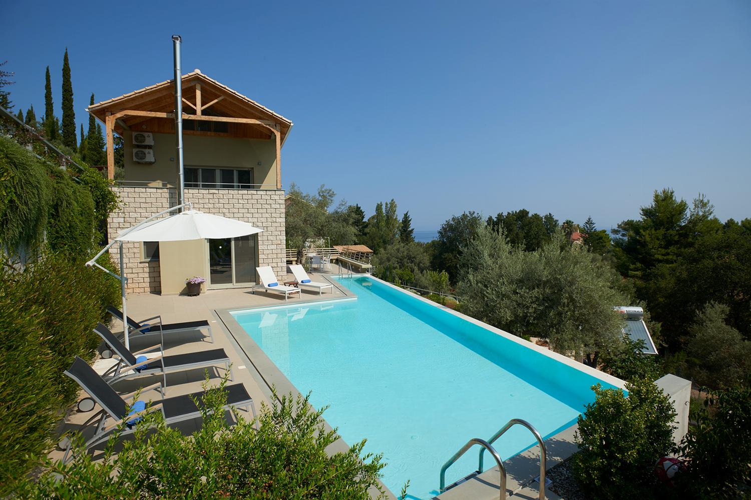 A patio view showcases lounge chairs lining a bright blue swimming pool next to a multi-story stone villa, offering a panoramic outlook toward the distant sea.