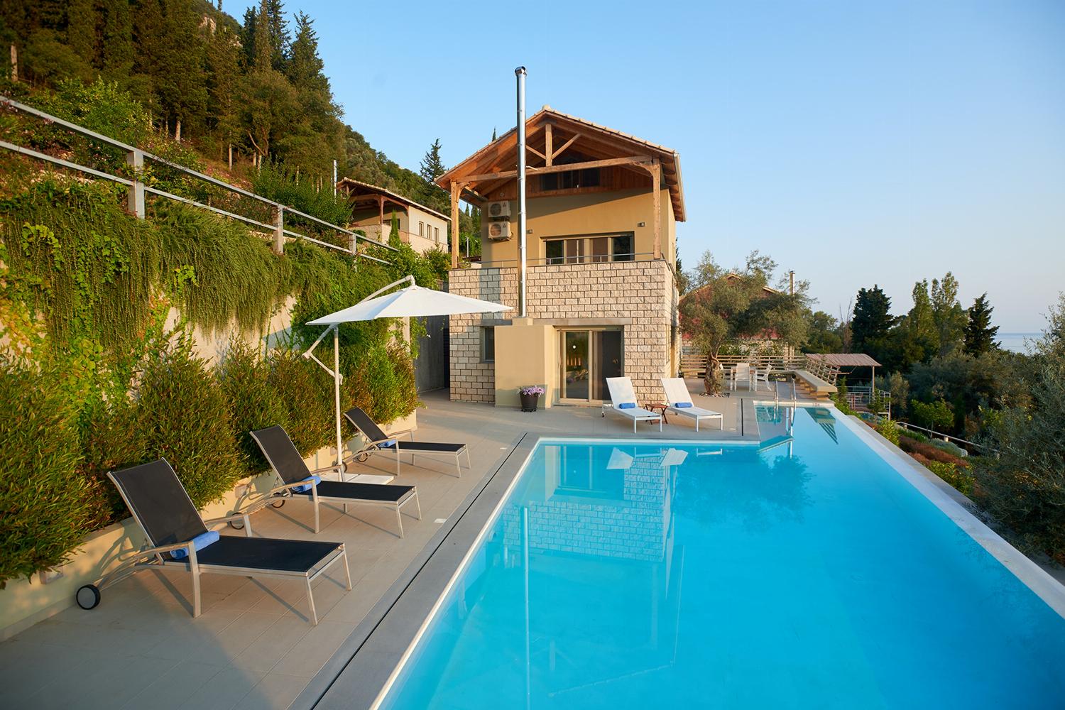 ,An elevated view showcases a multi-level stone villa nestled into a lush, green hillside, featuring an outdoor swimming pool, patio lounge chairs, and a dining terrace.