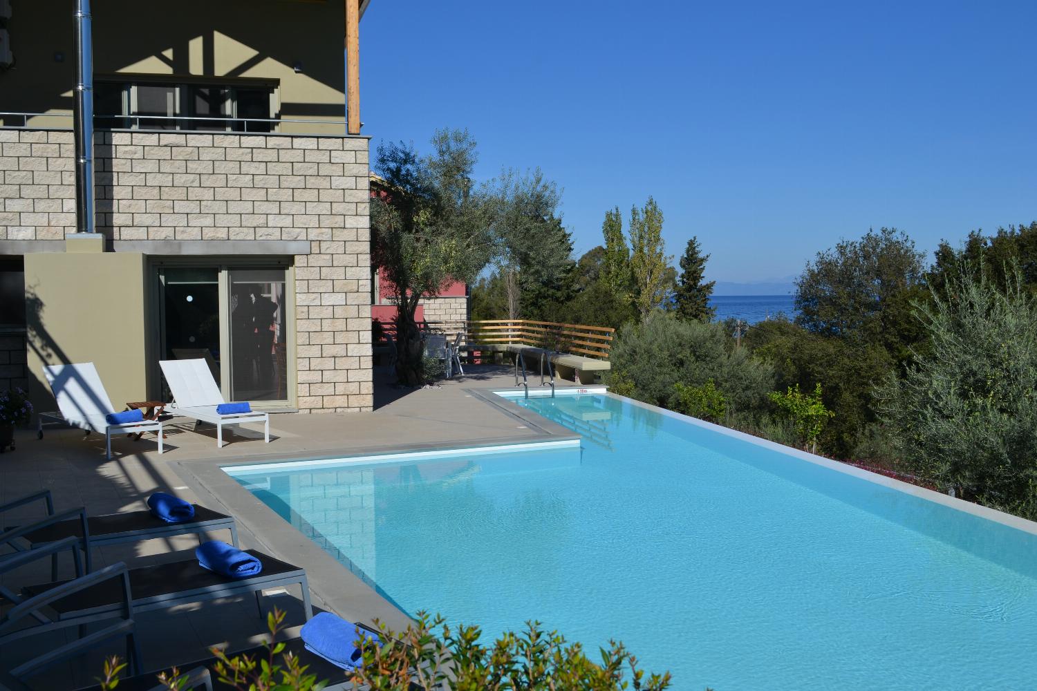 An outdoor swimming pool is bordered by lounge chairs and a stone villa patio, offering a sweeping view of green treetops and the sea beyond.
