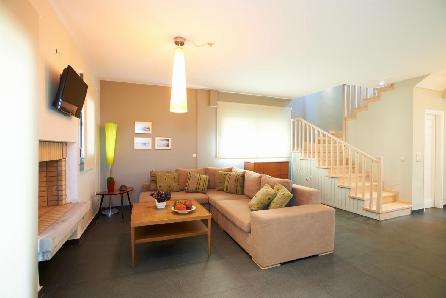 A spacious open-plan living room features a beige sectional sofa with red accent pillows, a wooden coffee table, a staircase, and a dining area in the background.