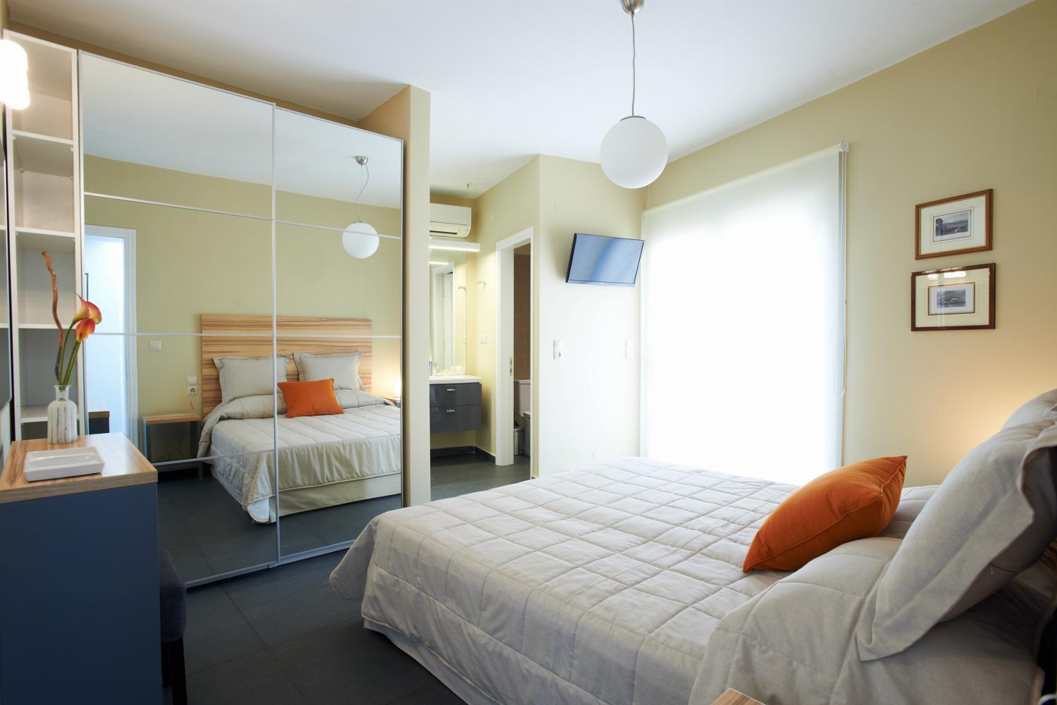 A bright bedroom features a double bed with a wooden headboard, a large mirrored wardrobe, and glass doors opening to a private outdoor seating area.