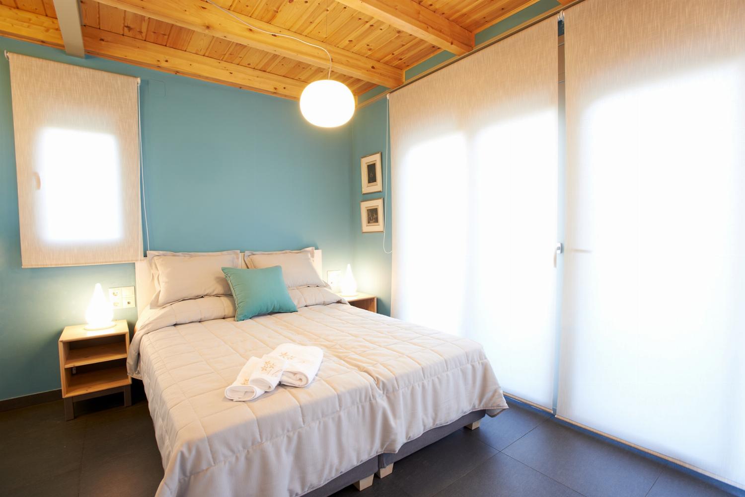 A charming bedroom features a double bed under an exposed wooden ceiling, blue accent walls, and wide glass doors that open to a private balcony with a mountain view.