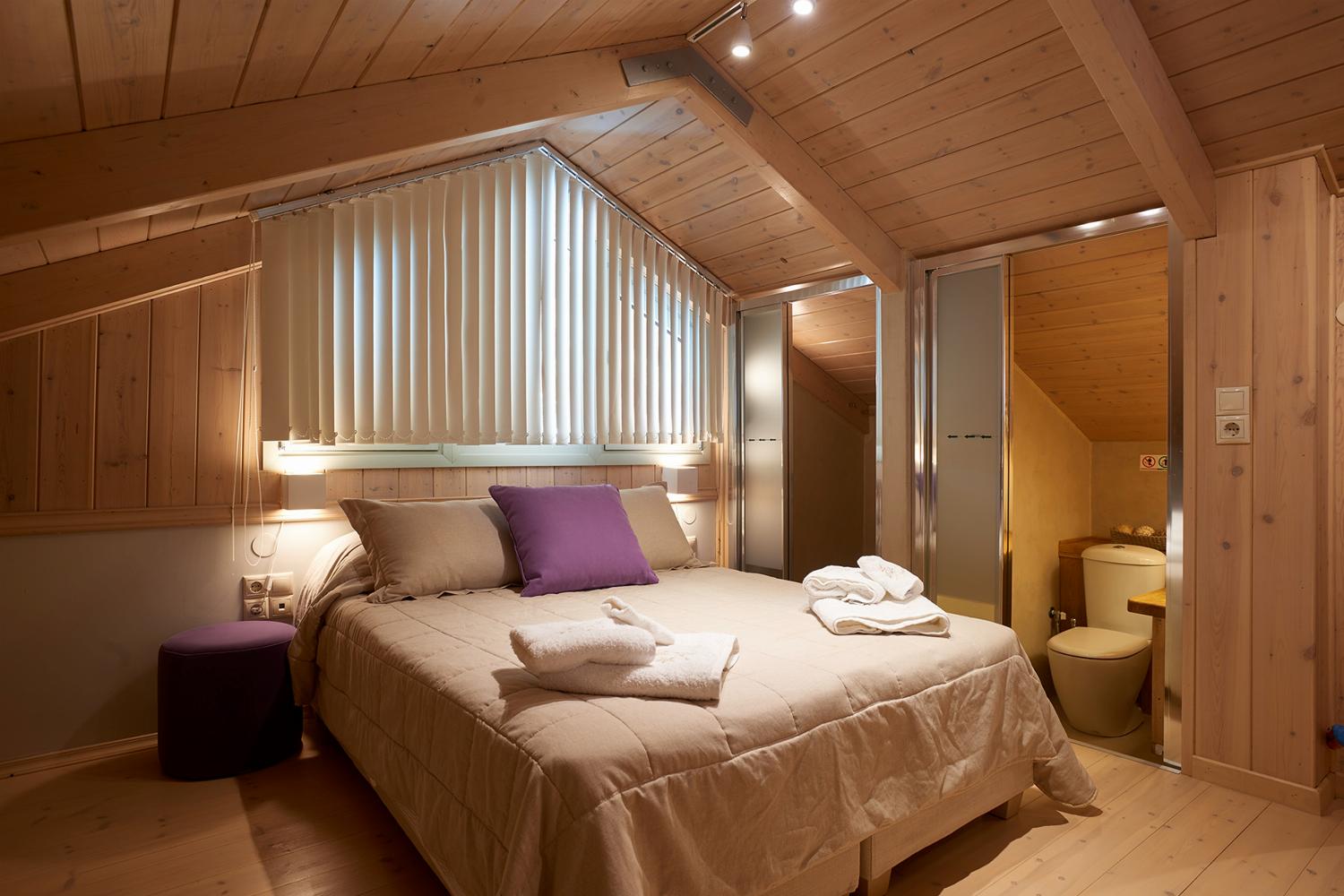 A rustic attic bedroom features wood-paneled walls and ceilings, a double bed with purple accent pillows, and tracking lights illuminating the sloped space.