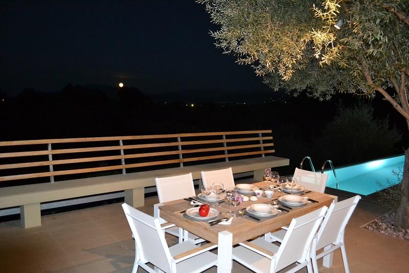 A wooden outdoor dining table is set with wine glasses and a lit candle on a terrace, offering a scenic view of the surrounding trees and sea under a pink sunset sky.