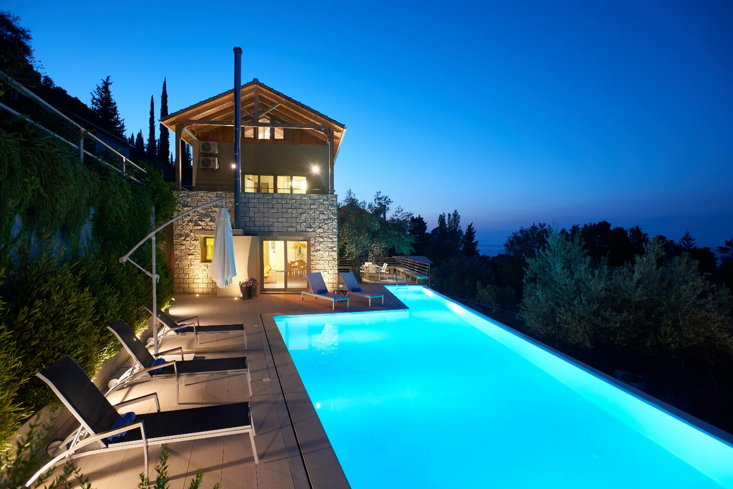 An illuminated outdoor swimming pool glows at dusk next to a multi-story stone villa, offering a panoramic view of the surrounding trees under a vibrant pink and purple sky.