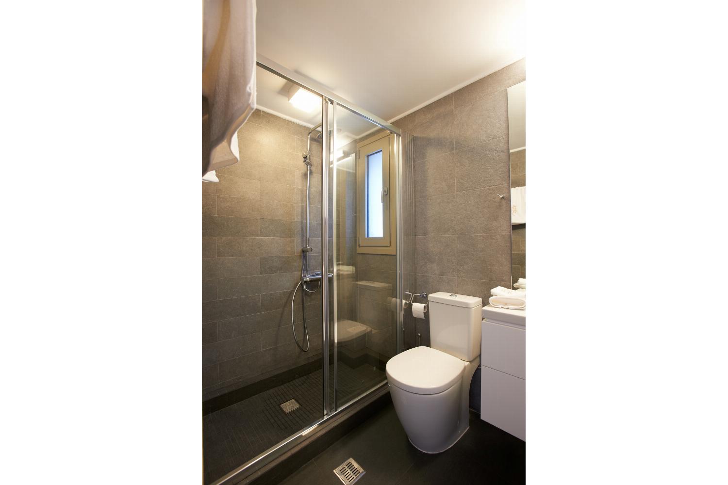 A contemporary bathroom features grey wall tiles, a glass-enclosed walk-in shower, a white vanity with drawers, and a large mirror reflecting an adjacent bedroom.