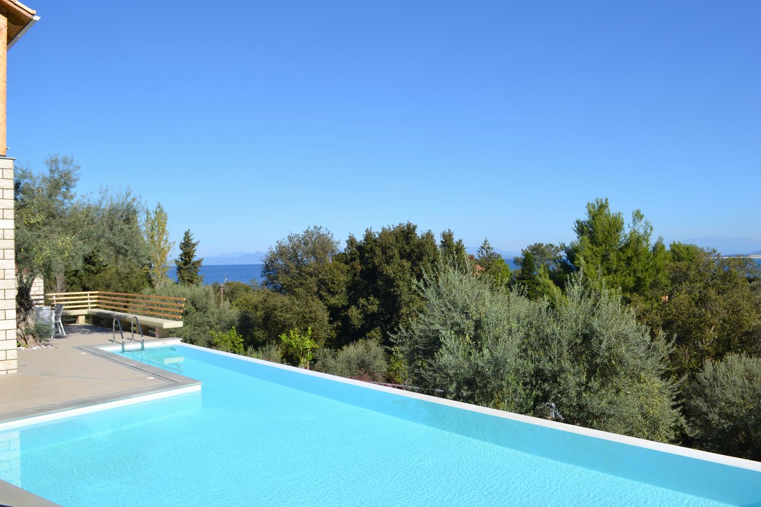 An expansive outdoor swimming pool is surrounded by lounge chairs on a spacious tiled deck, overlooking a vast landscape of green trees under a bright blue sky.