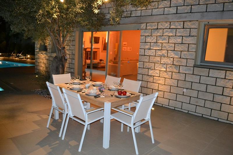A wooden outdoor dining table sits on a sun-drenched terrace next to a stone villa, featuring a private swimming pool and a lush mountain landscape in the background.
